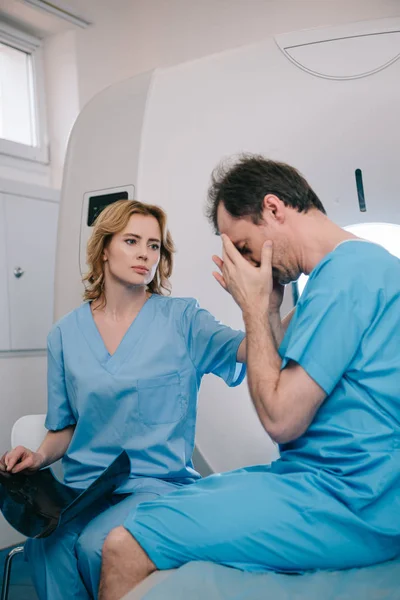 Hombre molesto cubriendo la cara con las manos mientras está sentado cerca del radiólogo en el hospital - foto de stock