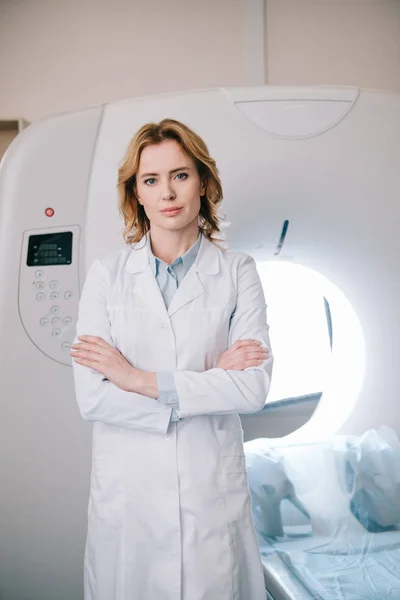 Schöner Radiologe, der mit verschränkten Armen neben dem Scanner steht und in die Kamera schaut — Stockfoto