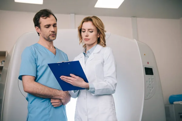Dos radiólogos de pie mirando el diagnóstico en el portapapeles mientras están de pie cerca del escáner de tomografía computarizada - foto de stock