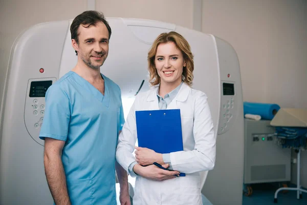 Aux médecins souriants regardant la caméra tout en se tenant près du scanner de tomographie informatique — Photo de stock