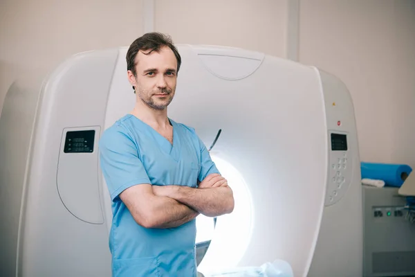 Lächelnder Radiologe, der mit verschränkten Armen neben einem Computertomographen steht und in die Kamera blickt — Stockfoto