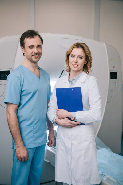 Красивый рентгенолог держит планшет, стоя рядом с ассистентом и глядя в камеру — стоковое фото