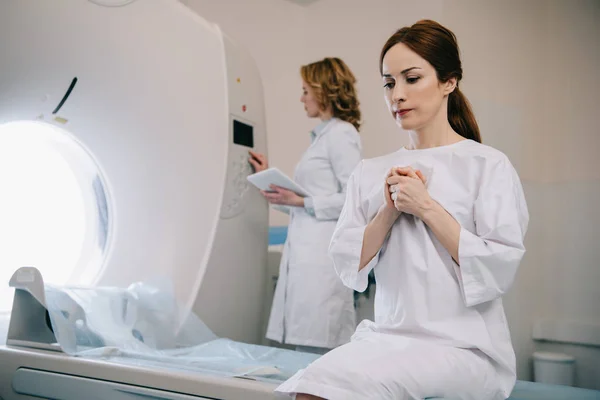 Enfoque selectivo de la mujer preocupada rezando mientras está sentada en la cama del escáner ct cerca del radiólogo - foto de stock