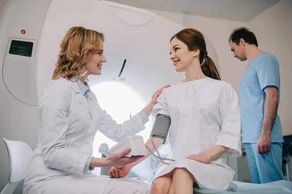 Médecin souriant mesurant la pression artérielle des patients avec tonomètre tout en assistant d'ajustement ct scanner — Stock Photo