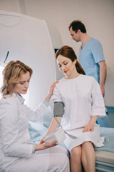 Selektiver Fokus des aufmerksamen Arztes, der den Blutdruck des Patienten mit einem Tonometer misst, während der Radiologe den ct-Scanner einstellt — Stockfoto