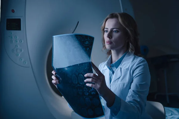 Beautiful doctor looking at x-ray diagnosis while sitting near computed tomography scanner — Stock Photo