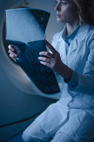 Médico reflexivo que sostiene el diagnóstico de rayos X mientras está sentado en el escáner de tomografía computarizada - foto de stock