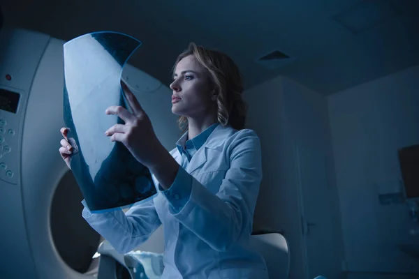 Beautiful thoughtful doctor looking at x-ray diagnosis while sitting near computed tomography scanner — Stock Photo