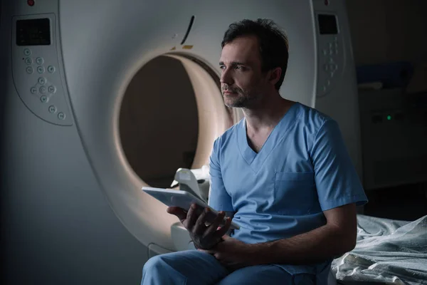 Serious radiologist holding digital tablet with x-ray diagnosis while sitting near computed tomography scanner and looking away — Stock Photo