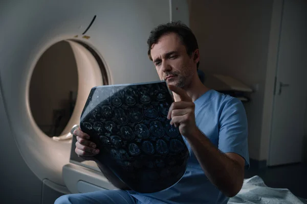 Médico atencioso olhando para o diagnóstico de tomografia enquanto sentado perto de tomógrafo — Fotografia de Stock