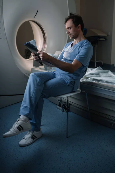 Thoughtful doctor examining x-ray diagnosis while sitting near computed tomography scanner — Stock Photo