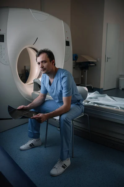 Серьезный рентгенолог, проводящий рентгеновскую диагностику, сидя рядом с компьютерным томографом и глядя в камеру — стоковое фото
