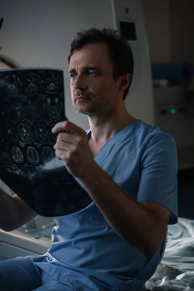 Серьезный врач, осматривающий рентгеновскую диагностику, сидя рядом со сканером КТ в больнице — стоковое фото
