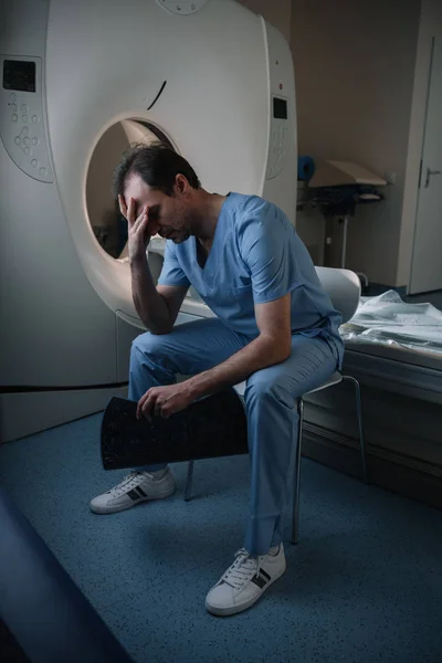 Radiologista exausto segurando o diagnóstico de raios-x enquanto sentado perto do tomógrafo no hospital — Fotografia de Stock