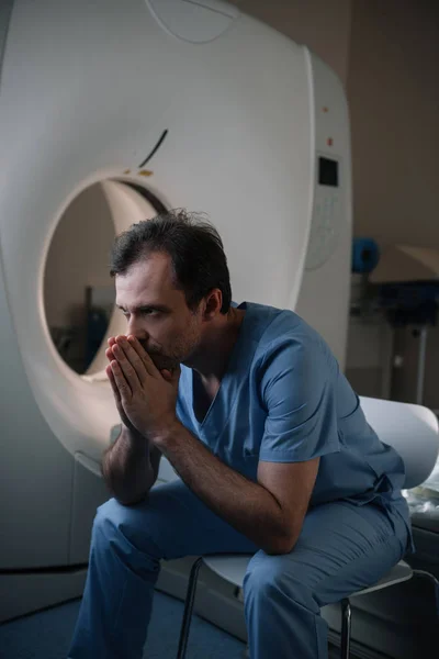 Thoughtful radiologist sitting near computed tomography scanner in hospital and looking away — Stock Photo