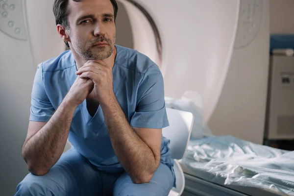 Pensive doctor sitting near computed tomography scanner in hospital and looking at camera — Stock Photo