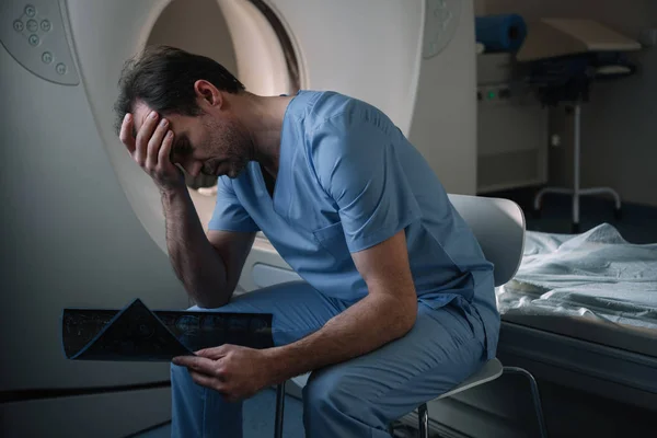 Exhausto sentado cerca del escáner de tomografía computarizada en el hospital y con diagnóstico de rayos X - foto de stock