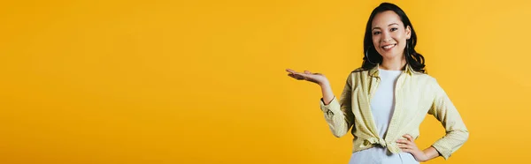 Feliz menina asiática apresentando algo isolado no amarelo — Fotografia de Stock