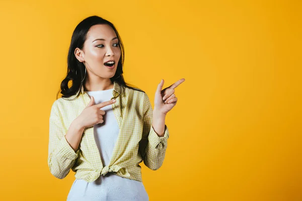 Fröhliches asiatisches Mädchen zeigt auf etwas Isoliertes auf gelb — Stockfoto