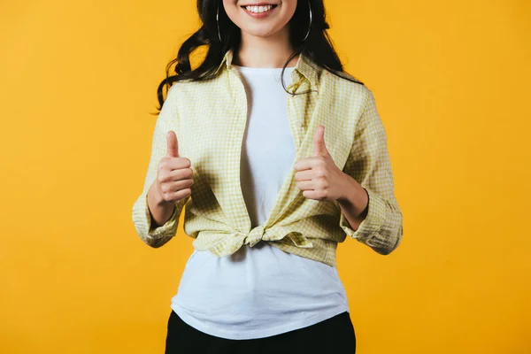 Обрізаний вид усміхненої дівчини, що показує великі пальці вгору ізольовано на жовтому — стокове фото