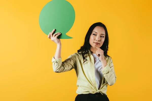 Вдумчивая азиатская женщина держит зеленый пузырь речи, изолированный на желтый — стоковое фото