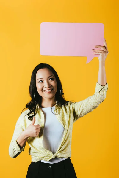 Mujer asiática casual mostrando el pulgar hacia arriba y sosteniendo la burbuja del habla rosa, aislado en amarillo - foto de stock