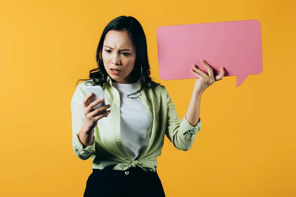 Impactado chica asiática usando teléfono inteligente y sosteniendo la burbuja del habla rosa, aislado en amarillo - foto de stock