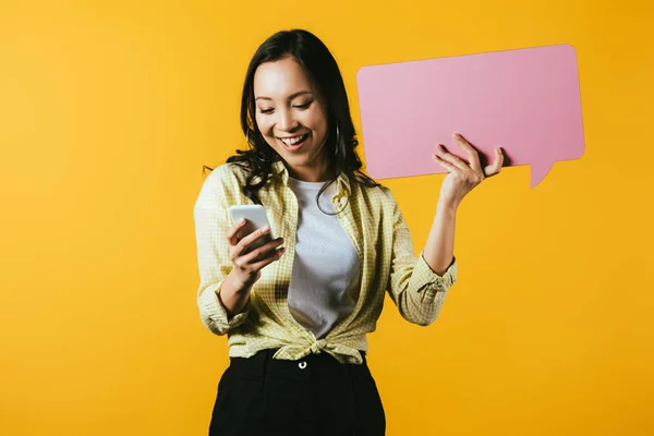 Casual asiático menina usando smartphone e segurando rosa discurso bolha, isolado no amarelo — Fotografia de Stock