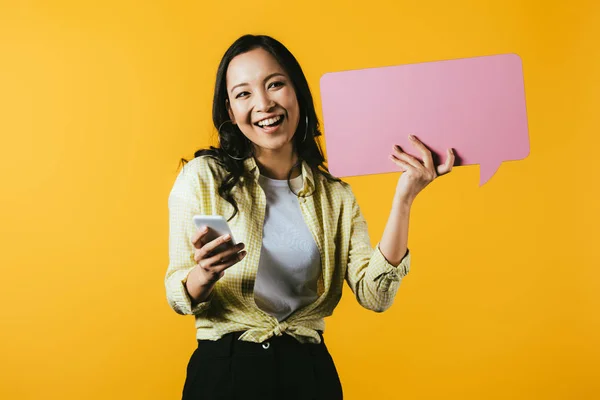 Счастливая азиатская девушка с помощью смартфона и проведение розовый пузырь речи, изолированные на желтый — стоковое фото
