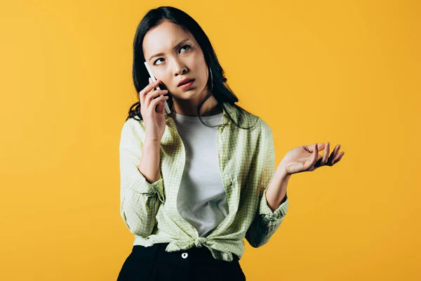 Confused asian girl talking on smartphone isolated on yellow — Stock Photo