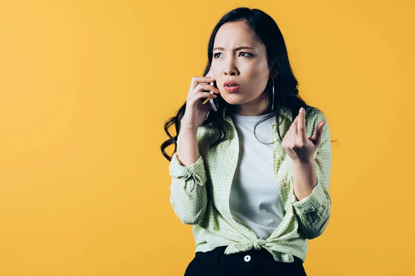 Worried asian young woman talking on smartphone isolated on yellow — Stock Photo