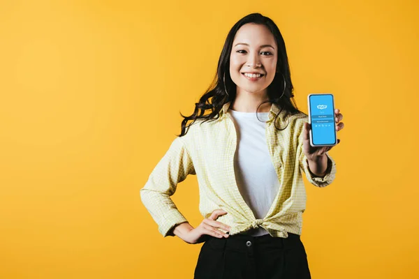 KYIV, UCRANIA - 16 DE ABRIL DE 2019: chica asiática sonriente mostrando teléfono inteligente con aplicación skype, aislado en amarillo - foto de stock