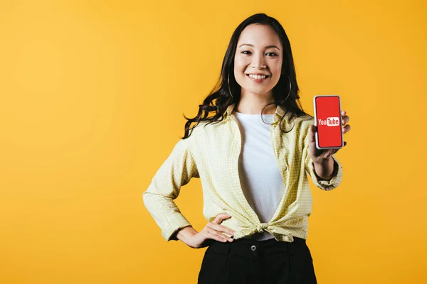 KYIV, UCRANIA - 16 DE ABRIL DE 2019: chica asiática sonriente mostrando teléfono inteligente con aplicación de youtube, aislado en amarillo - foto de stock