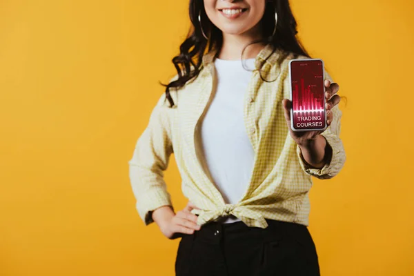 Обрізаний вид брюнетки дівчина показує смартфон з додатком торгових курсів, ізольований на жовтому — стокове фото