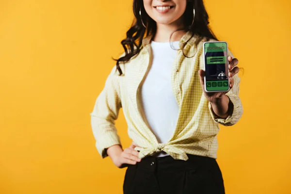 Vista recortada de la chica sonriente que muestra el teléfono inteligente con aplicación de reserva, aislado en amarillo - foto de stock