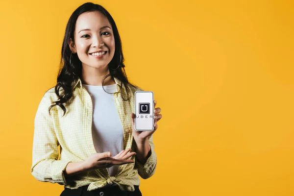 KYIV, UCRANIA - 16 DE ABRIL DE 2019: chica asiática sonriente presentando teléfono inteligente con aplicación uber, aislado en amarillo - foto de stock