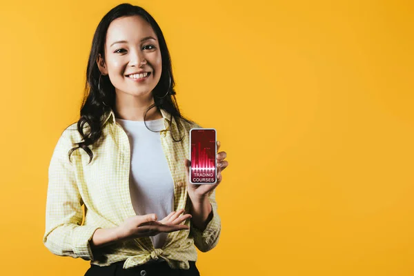Menina asiática feliz apresentando smartphone com cursos comerciais, isolado no amarelo — Fotografia de Stock