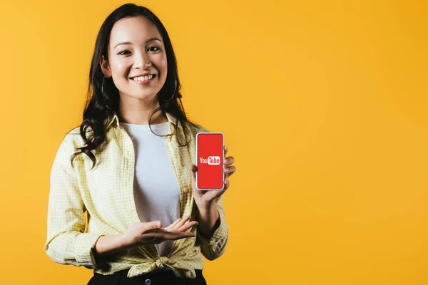 Kyiv, ukraine - 16. April 2019: lächelndes asiatisches Mädchen präsentiert Smartphone mit Youtube-App, isoliert auf gelb — Stockfoto