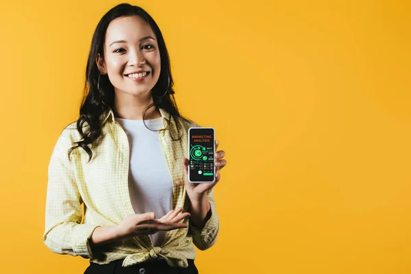 Chica asiática feliz presentando smartphone con análisis de marketing, aislado en amarillo - foto de stock