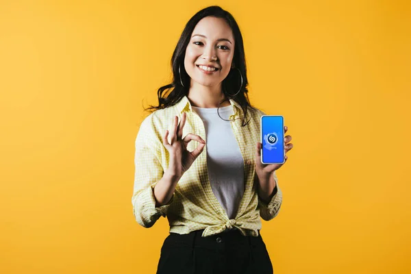 KYIV, UKRAINE - APRIL 16, 2019: smiling asian girl showing ok sign and smartphone with shazam app, isolated on yellow — Stock Photo