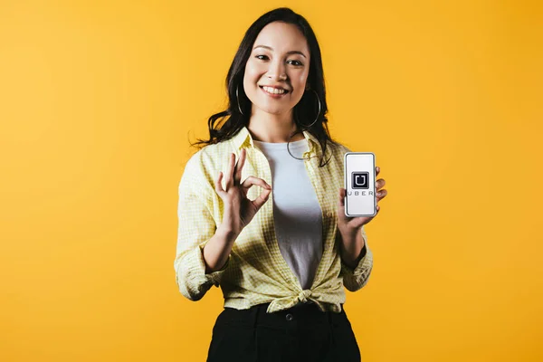 KYIV, UKRAINE - APRIL 16, 2019: smiling asian girl showing ok sign and smartphone with uber app, isolated on yellow — Stock Photo