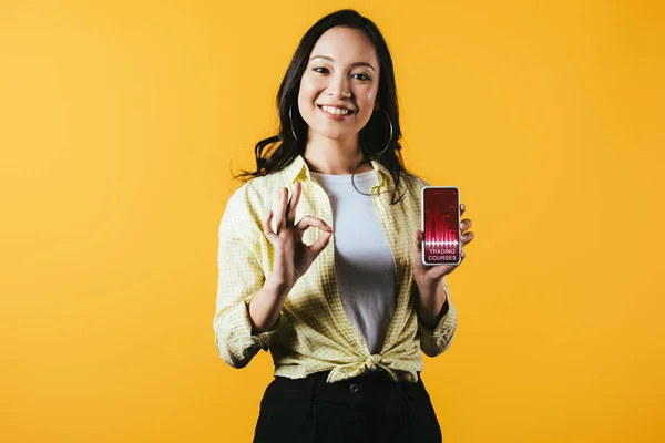 Счастливая азиатская девушка показывает ОК знак и смартфон с торговыми курсами, изолированные на желтый — стоковое фото
