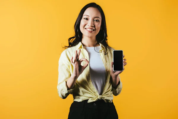 Attractive asian girl showing ok sign and smartphone with blank screen isolated on yellow — Stock Photo