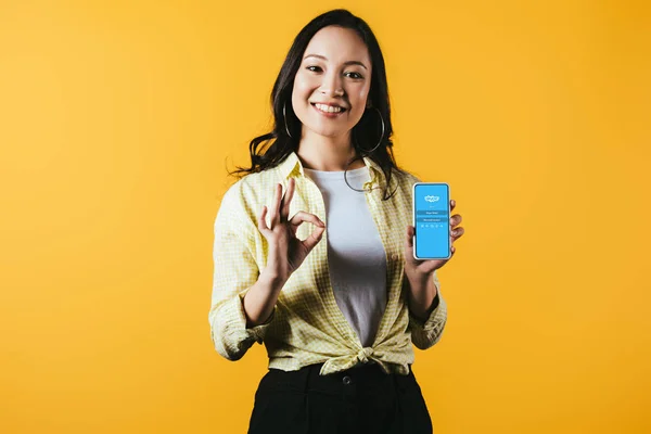 KYIV, UKRAINE - APRIL 16, 2019: smiling asian girl showing ok sign and smartphone with skype app, isolated on yellow — Stock Photo