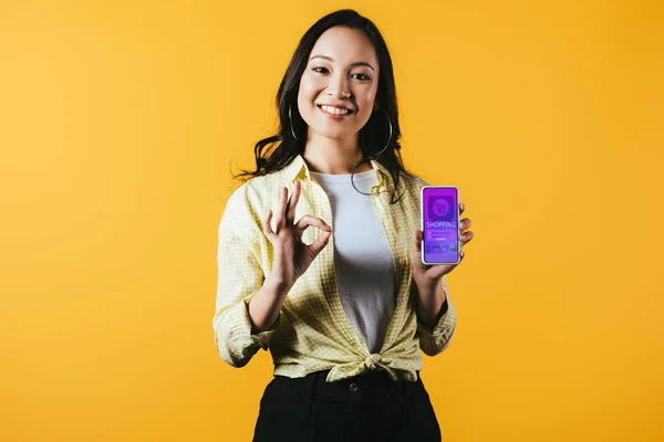 Улыбающаяся азиатская девушка показывает знак ОК и смартфон с приложением для покупок, изолированный на желтый — стоковое фото