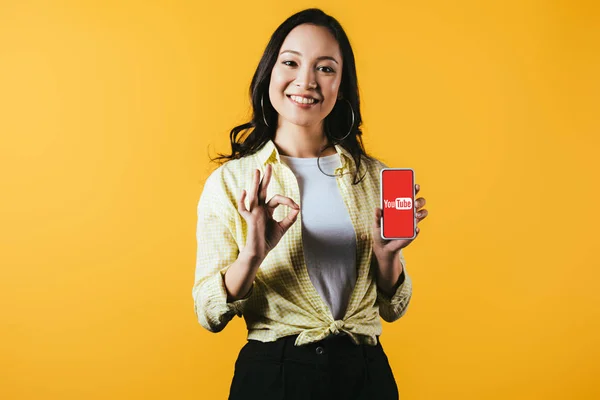 KYIV, UKRAINE - APRIL 16, 2019: smiling asian girl showing ok sign and smartphone with youtube app, isolated on yellow — Stock Photo