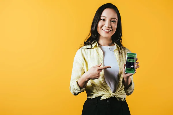 Sonriente chica asiática apuntando a teléfono inteligente con aplicación de reserva, aislado en amarillo - foto de stock