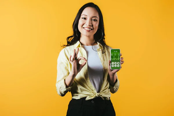 Sourire asiatique fille montrant ok signe et smartphone avec application de santé, isolé sur jaune — Photo de stock