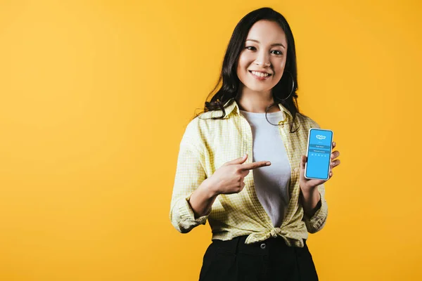 KYIV, UCRANIA - 16 DE ABRIL DE 2019: atractiva chica asiática sonriente apuntando al teléfono inteligente con aplicación skype, aislada en amarillo - foto de stock