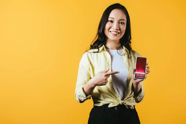 Lächelndes asiatisches Mädchen zeigt auf Smartphone mit Handelskursen, isoliert auf gelb — Stockfoto